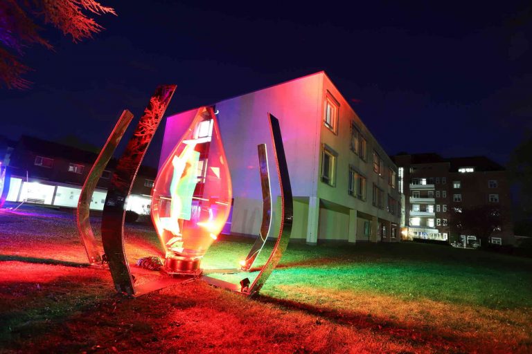 Colorful sculpture with lights at night near buildings.